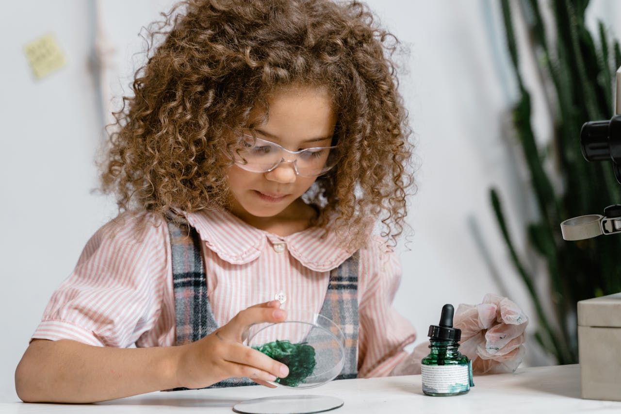 A young girl in glasses conducting a science experiment with green liquid indoors.
